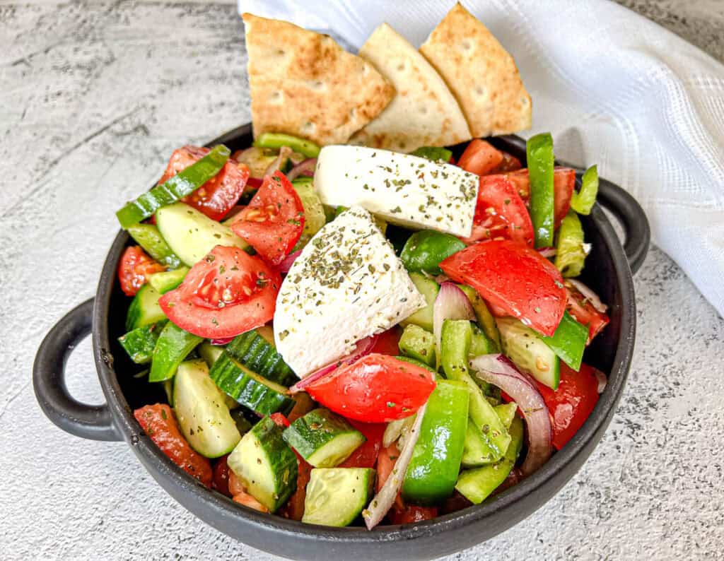 A bowl with Horiatiki Salad with feta cheese on top and pita bread on the side.