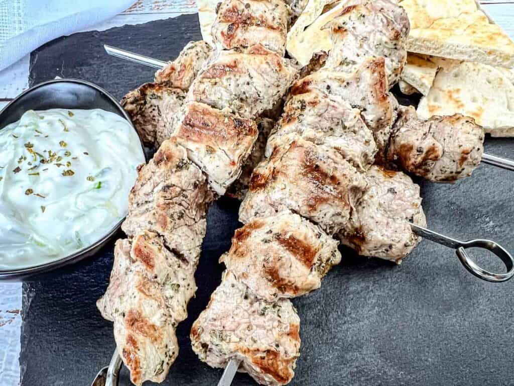 Close-up shot of Pork souvlaki on a slate plate with a sauce beside.