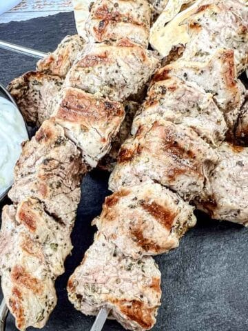 Four pieces of pork souvlaki with a bowl of white sauce and pieces of flatbread on a black slate serving board.