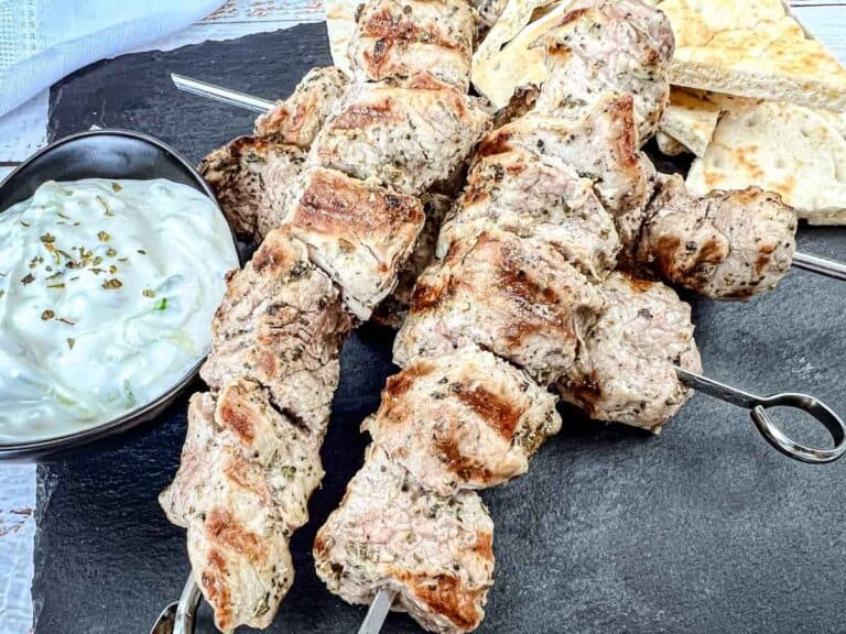 Four pieces of pork souvlaki with a bowl of white sauce and pieces of flatbread on a black slate serving board.