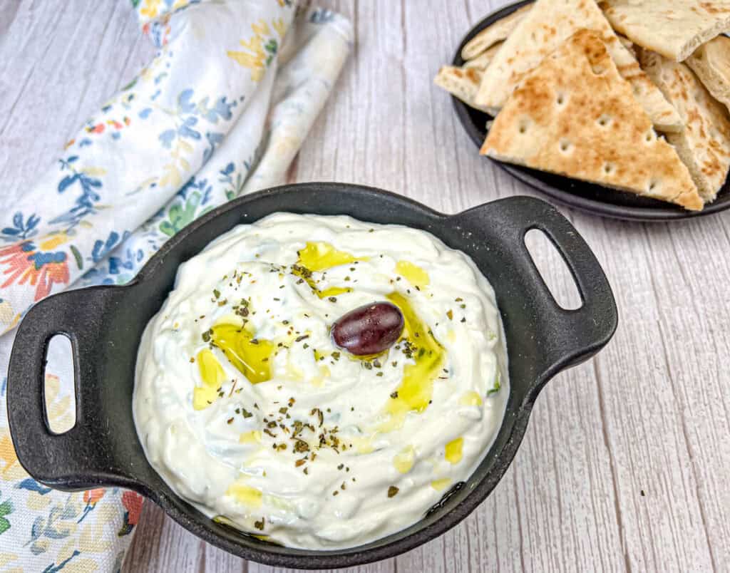 Tzatziki Sauce on a black bowl beside pita bread on a plate.