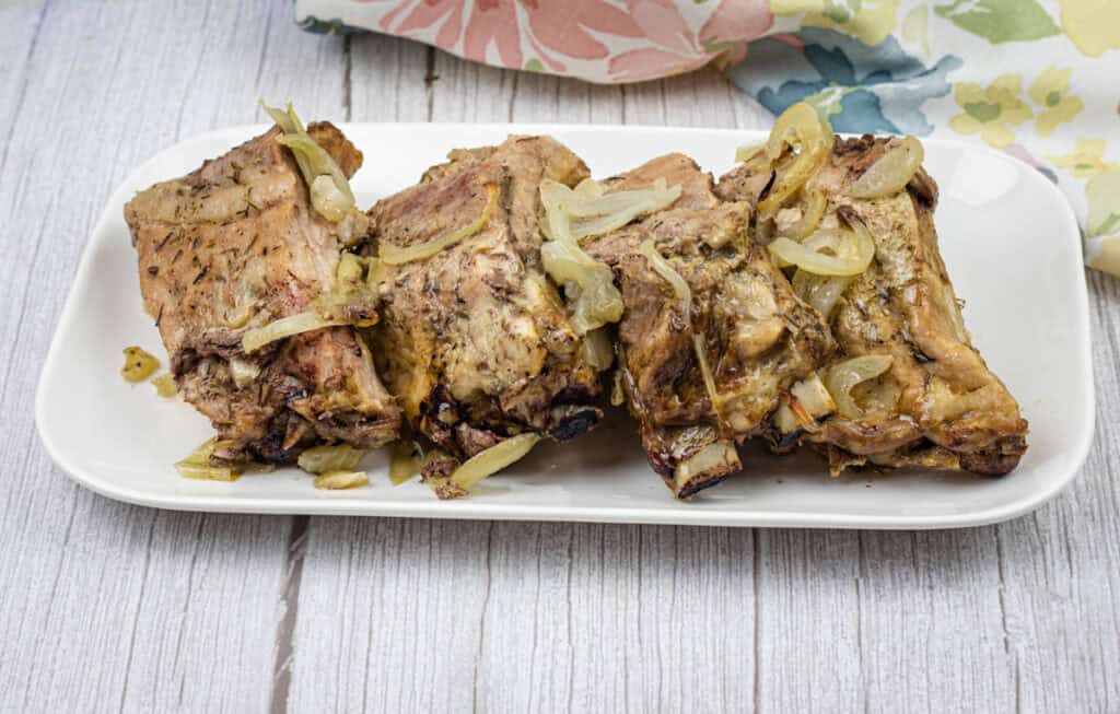 Oven-baked ribs with onion and garlic on top served on a white rectangular plate.