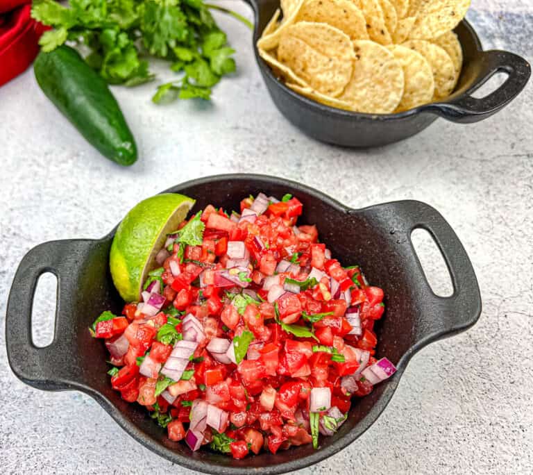 Pico de gallo in a black cast iron bowl with chips and jalapeño in the background.