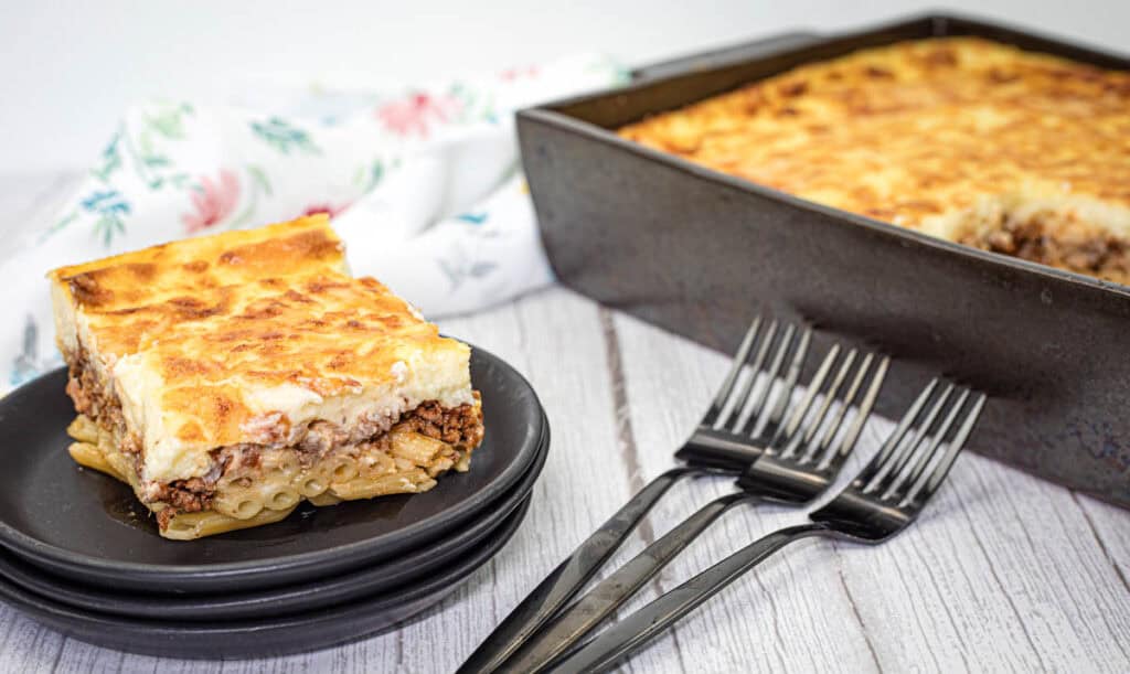 A slice of Pastitsio on a black plate beside the tray of Pastitsio.