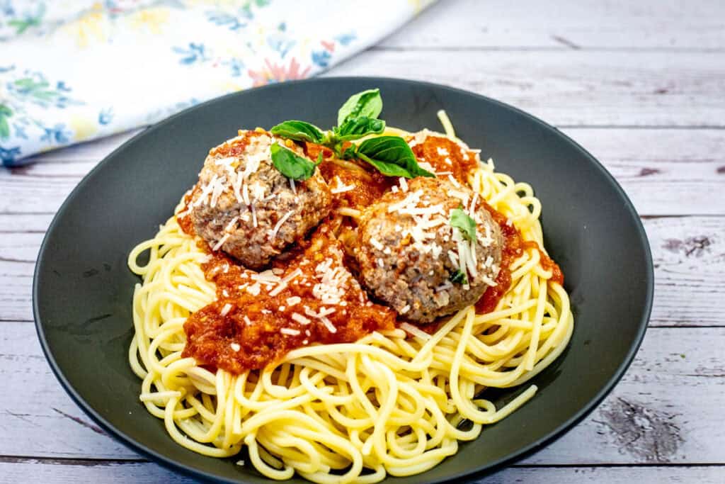 A plate of spaghetti topped with tomato sauce, two large meatballs, grated cheese, and a sprig of basil.