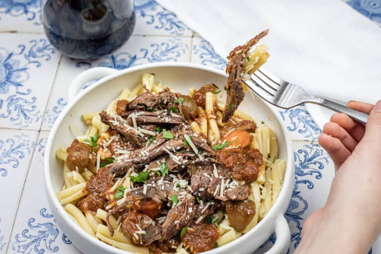 A hand lifting a fork with a bite of Italian pot roast pasta from a bowl.