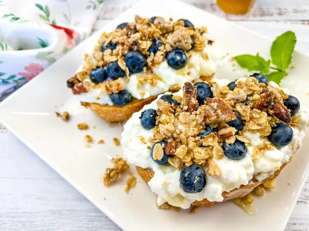 A plate of cottage cheese toast with blueberries, next to a honey dipper.