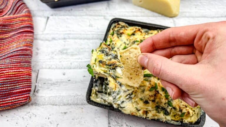Hand holding a chip with spinach artichoke dip over a square dish of the same dip.