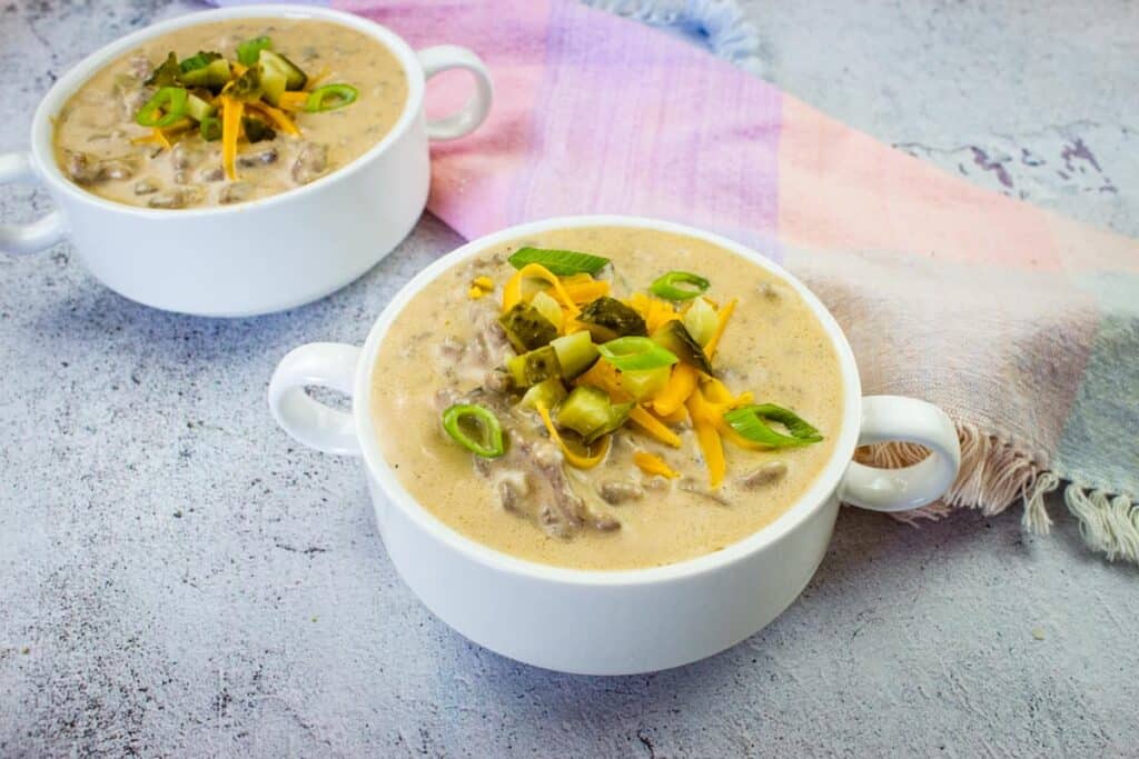 Two bowls of creamy soup topped with chopped green onions, shredded cheese, and diced pickles, placed on a textured surface with a pastel cloth in the background.