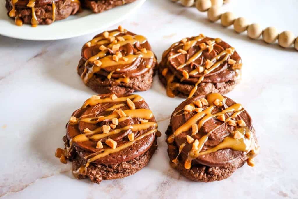 Chocolate Caramel Cookies.Chocolate Caramel Cookies with frosting, caramel drizzle, and chopped nuts on a white marble surface.