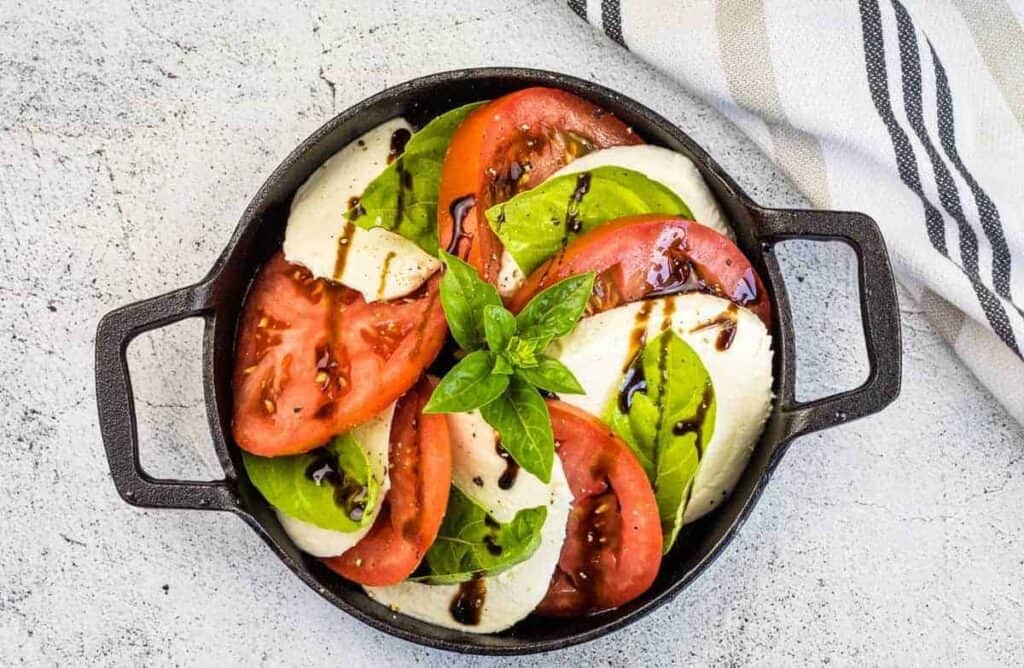 A caprese salad with sliced tomatoes, fresh mozzarella, and basil leaves, drizzled with balsamic glaze, served in a black dish on a light textured surface. A striped cloth is nearby.