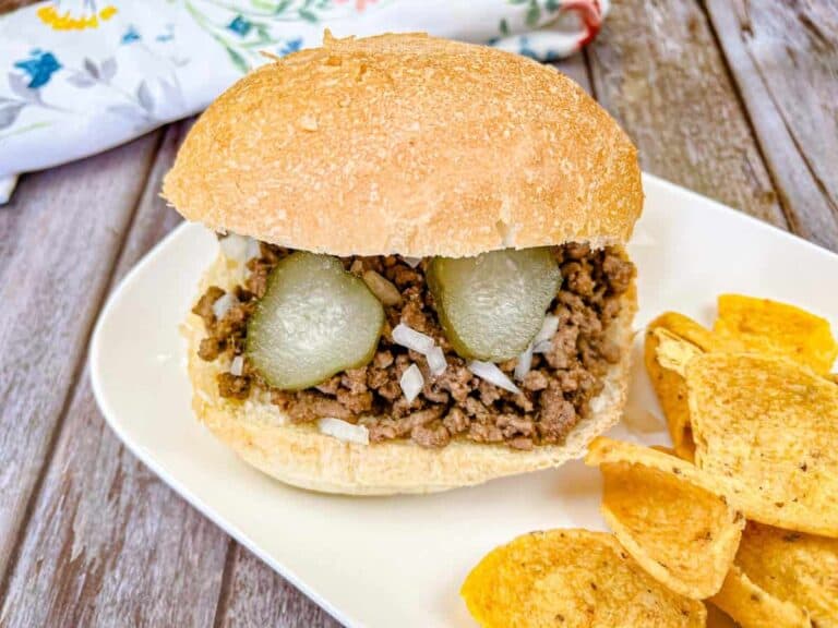 A sloppy joe sandwich with ground beef, pickles, and onions is on a plate next to a serving of potato chips. A floral-patterned napkin is in the background.