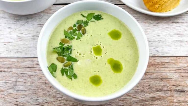 Bowl of green soup garnished with pumpkin seeds, parsley, and small oil drizzles on a wooden table next to a partial view of bread.