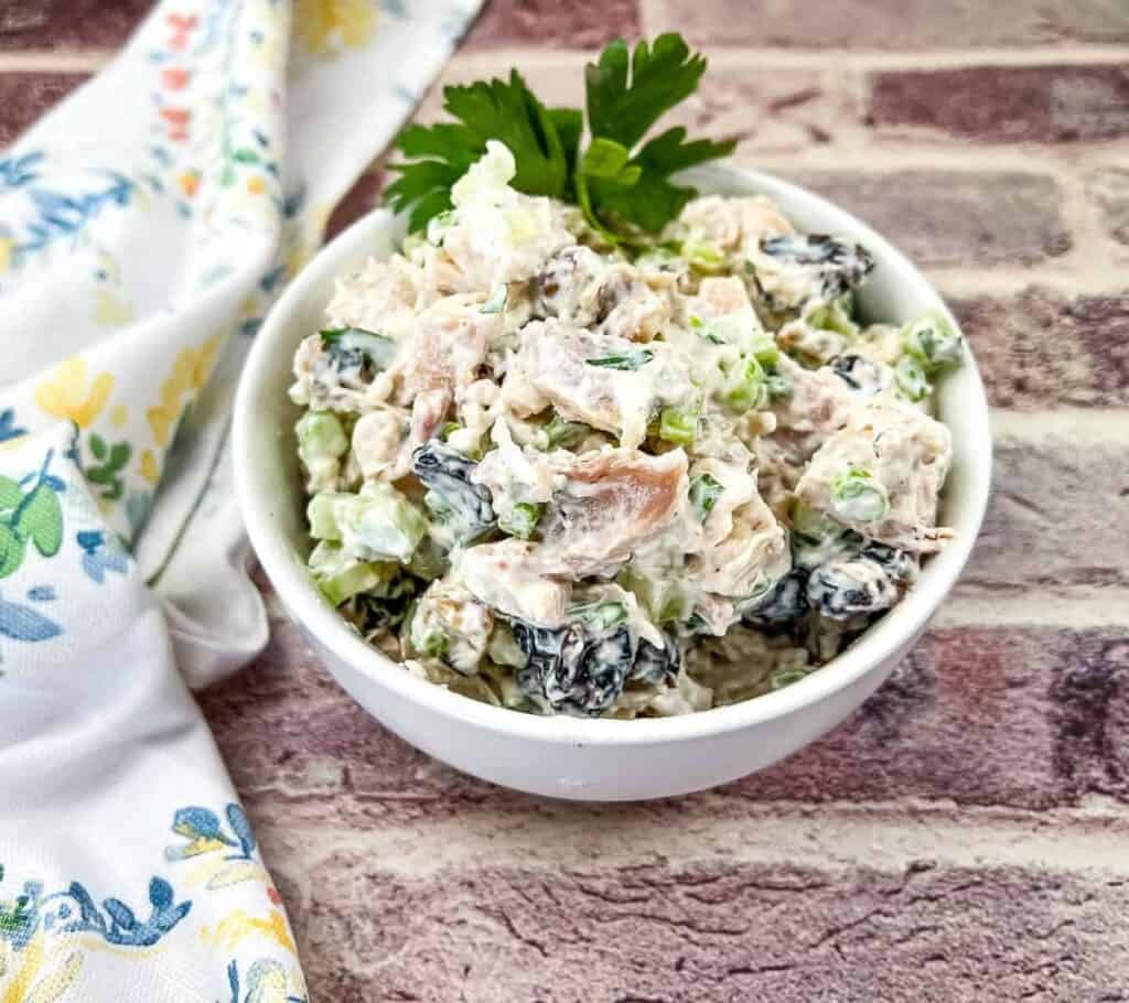 A bowl of chicken salad with chopped greens and black olives, garnished with parsley, placed on a brick-textured surface next to a floral-patterned cloth.