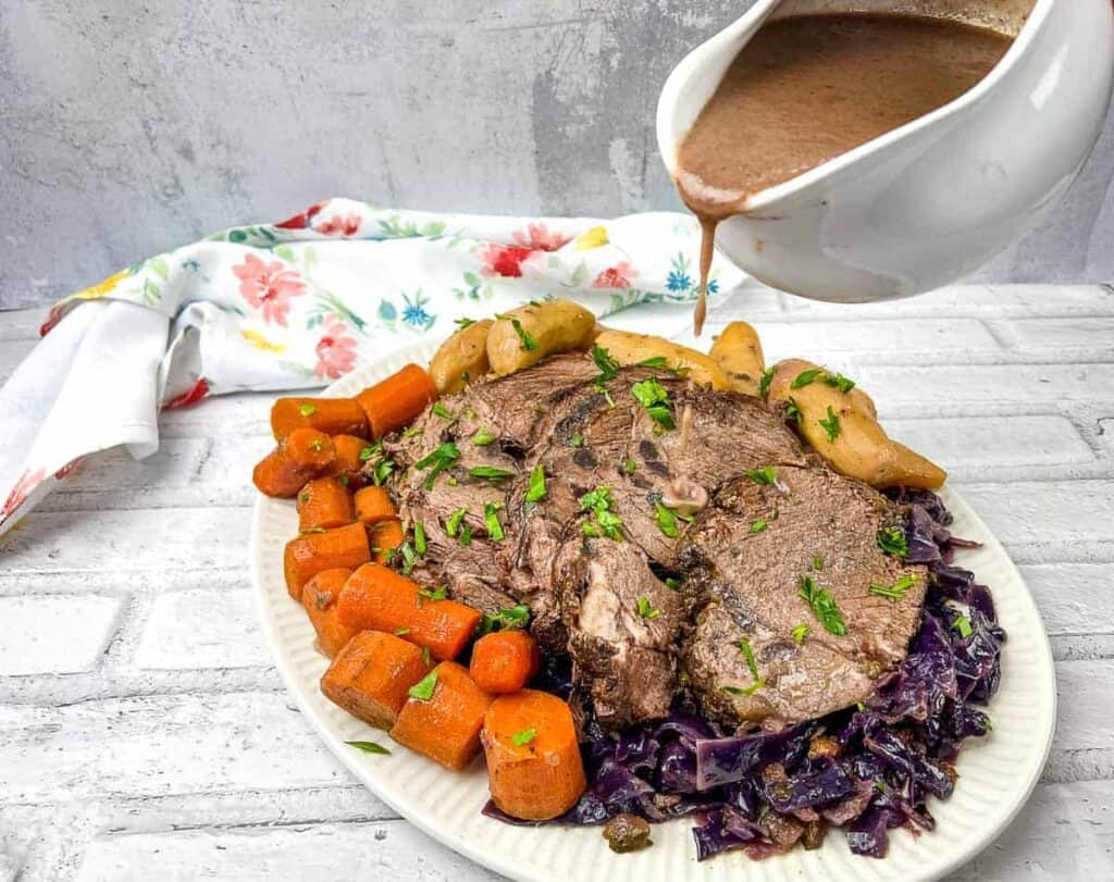 A plate of sliced roast beef with carrots, potatoes, and red cabbage. Gravy is being poured from a white gravy boat. A floral napkin is in the background.