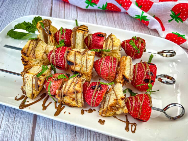 Skewers with grilled cake cubes and strawberries drizzled with chocolate and garnished with mint on a white plate.