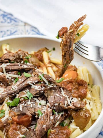 A hand holds a fork with Italian Pot Roast Pasta in a white bowl, topped with grated cheese and herbs.
