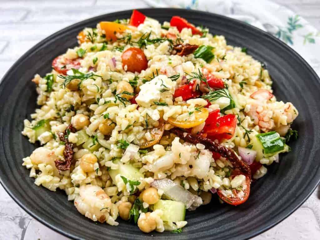 A plate of mixed salad featuring bulgur, chickpeas, cherry tomatoes, cucumbers, red onions, feta cheese, and herbs, served on a black dish.
