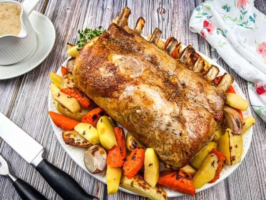 Roasted rack of lamb on a platter with assorted vegetables, including potatoes and carrots, on a wooden table. A knife, fork, and gravy boat are nearby.