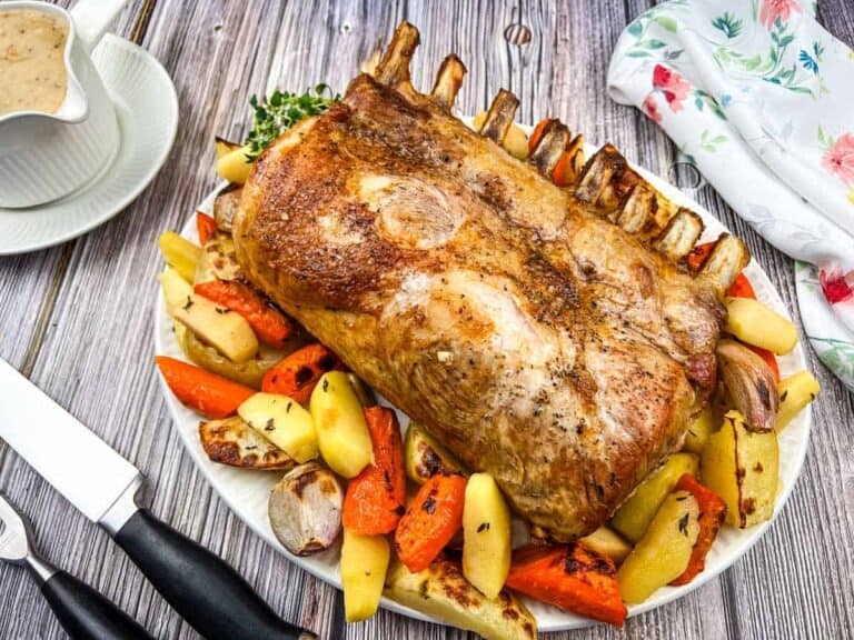 Roasted rack of lamb on a platter with assorted vegetables, including potatoes and carrots, on a wooden table. A knife, fork, and gravy boat are nearby.