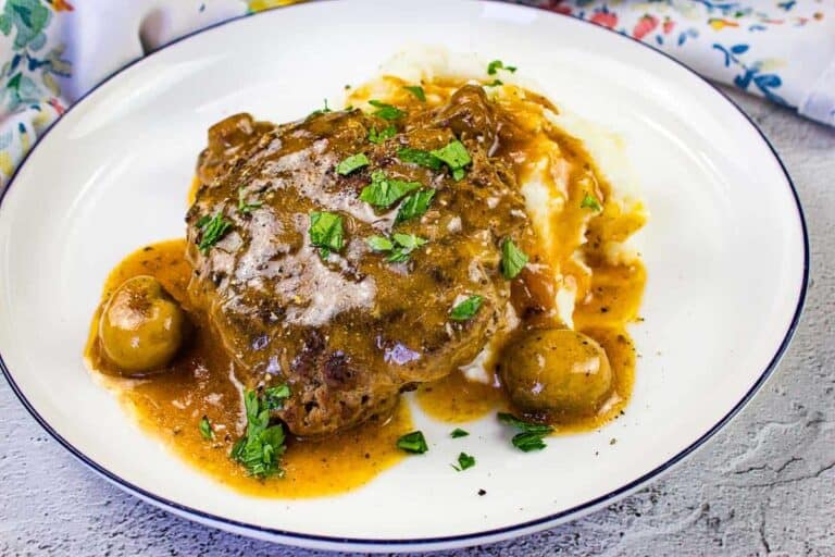 A plate with Salisbury steak topped with brown gravy, served with mashed potatoes and mushrooms, garnished with chopped parsley.
