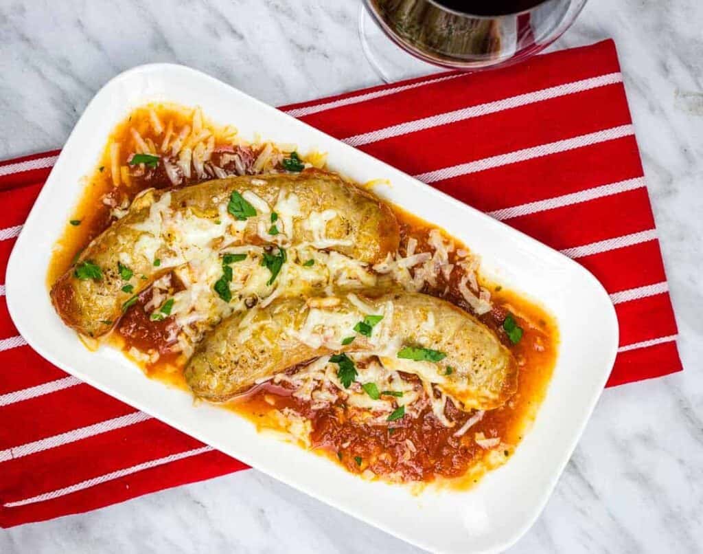 Two sausages topped with cheese and herbs on tomato sauce, served on a white plate over a red striped napkin, next to a glass of red wine.