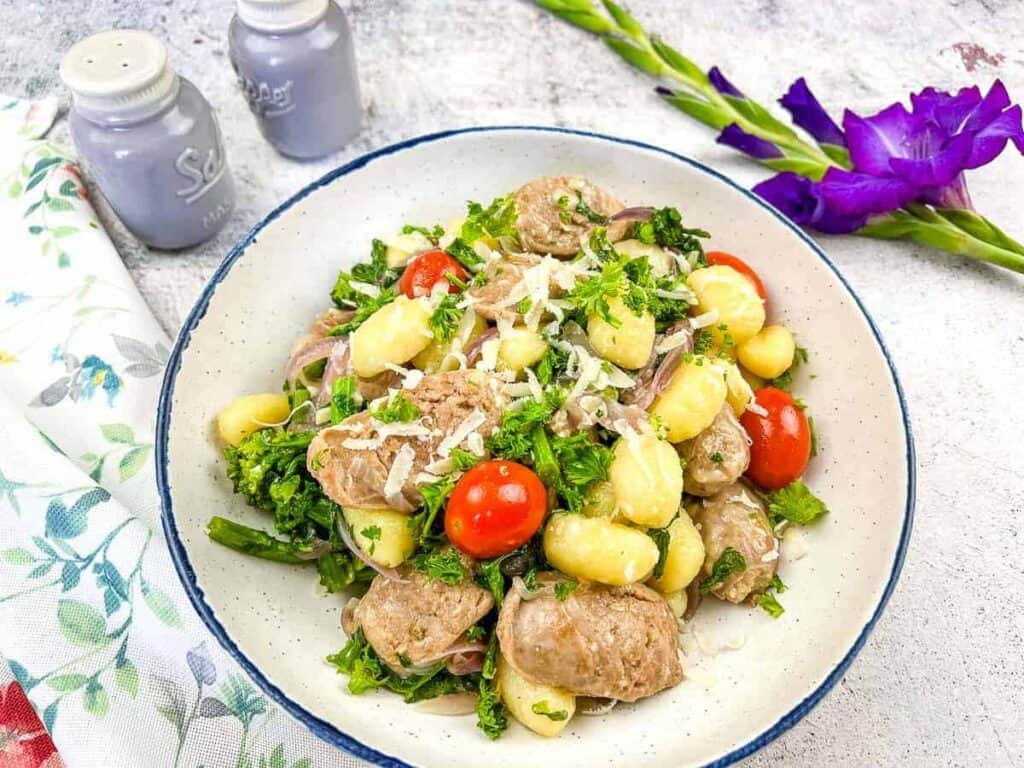 A bowl of gnocchi with sausage, broccoli, cherry tomatoes, and grated cheese. A floral napkin, salt and pepper shakers, and a purple flower are in the background.