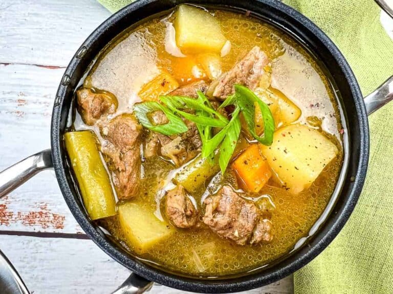 Soup with meat, potatoes, carrots, and green onion slices in a black pot on a wooden surface with a green cloth napkin.
