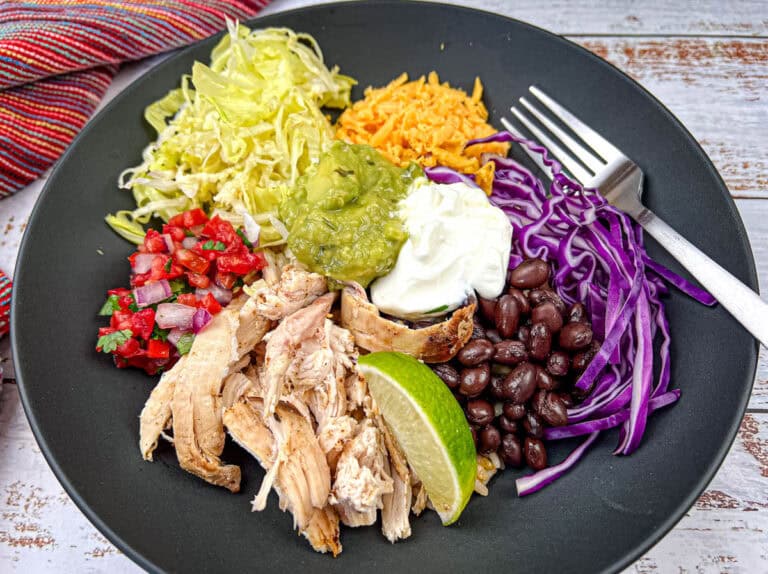 A black plate with shredded chicken, cabbage, red cabbage, black beans, shredded cheese, guacamole, sour cream, salsa, and a lime wedge, arranged next to a fork.