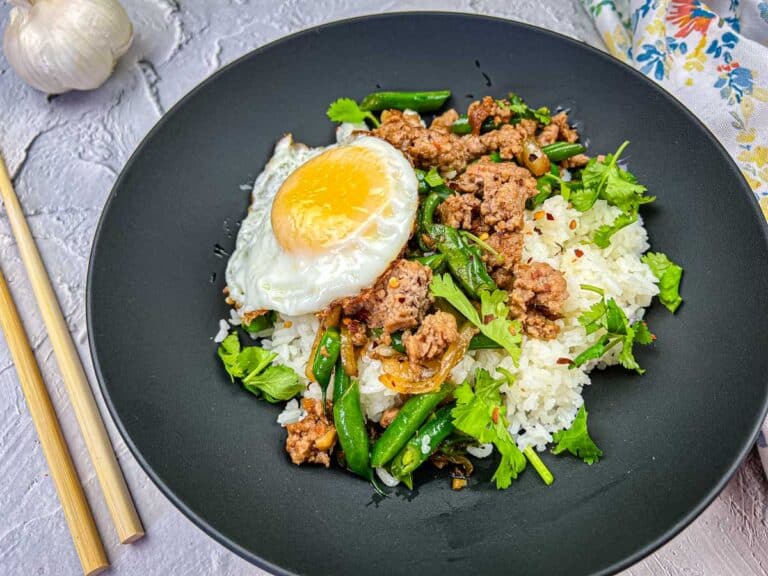 A black plate with rice topped with minced meat, a sunny-side-up egg, green beans, herbs, and spices. Chopsticks are placed beside the plate.