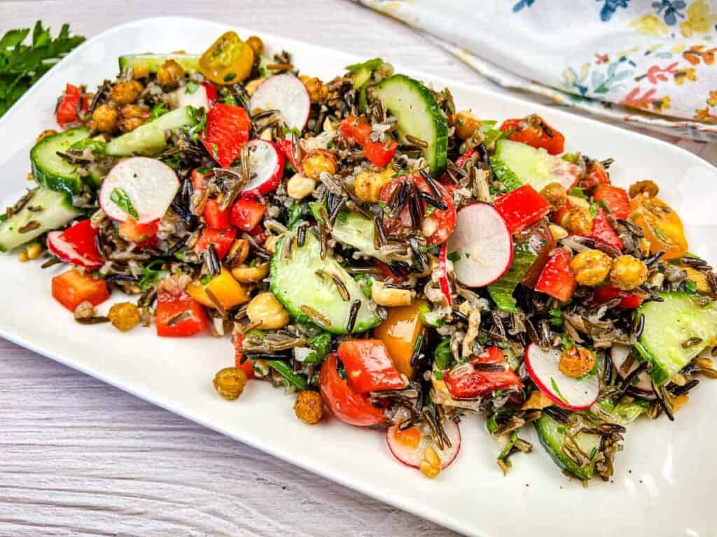 A colorful salad with wild rice, sliced cucumbers, radishes, cherry tomatoes, red bell peppers, and chickpeas on a rectangular white plate.