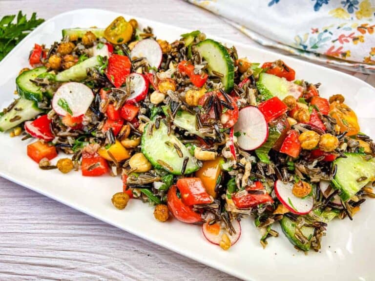A colorful salad with wild rice, sliced cucumbers, radishes, cherry tomatoes, red bell peppers, and chickpeas on a rectangular white plate.