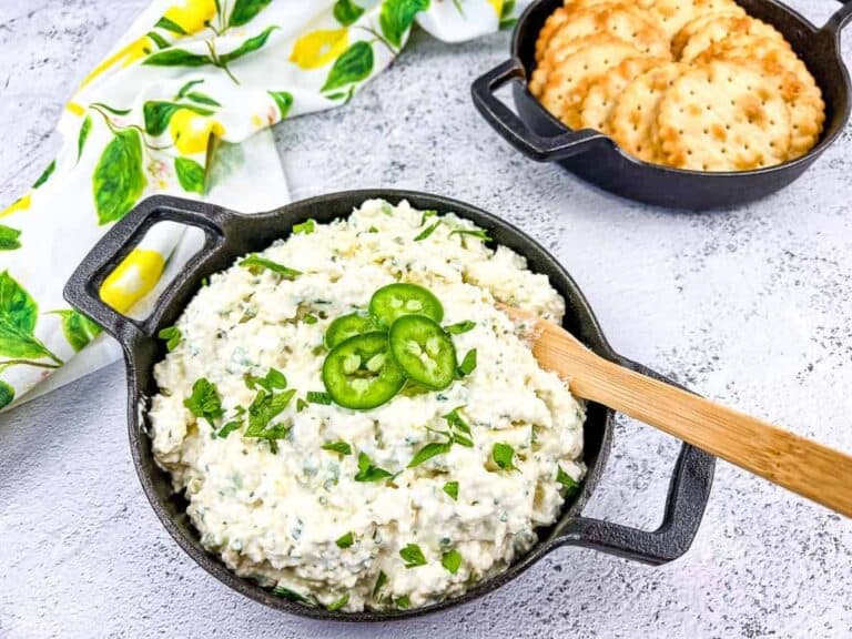 A bowl of jalapeño dip garnished with sliced jalapeños and herbs, with a wooden spoon. Crackers are in a nearby dish. A lemon-patterned cloth is partially visible.