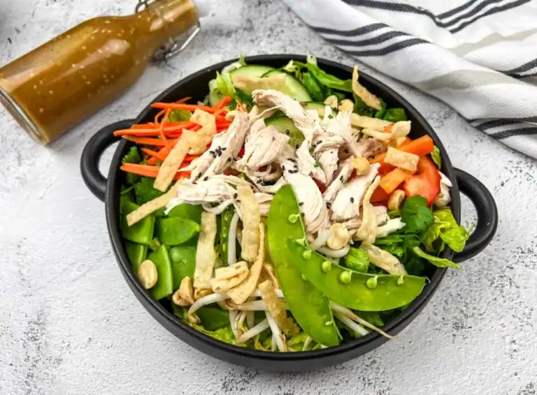 Bowl of salad with sliced chicken, snap peas, carrot strips, bean sprouts, and cashews. Dressing in a glass bottle nearby on a white surface with a striped napkin.