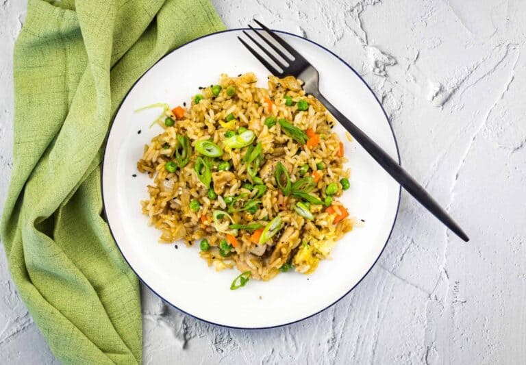 A plate of fried rice with vegetables and garnished with green onions, placed on a white plate with a black fork and a green cloth beside it.