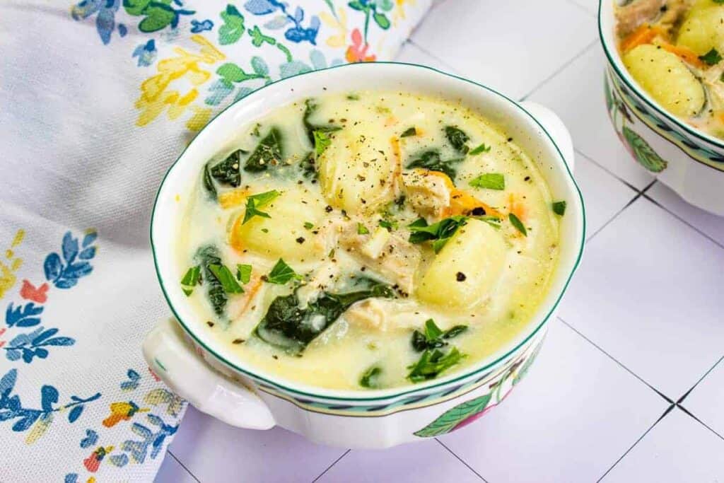 A bowl of creamy soup with gnocchi, spinach, shredded chicken, and herbs, placed on a floral-patterned tablecloth.