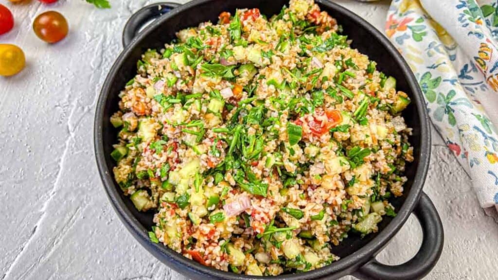 A bowl of tabbouleh with chopped parsley, bulgur, tomatoes, cucumber, and onions on a textured white surface.