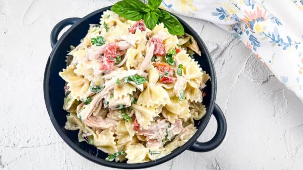 A black bowl of bowtie pasta salad with creamy dressing, red bell peppers, shredded chicken, and fresh greens, garnished with a basil leaf. A floral napkin is beside the bowl.