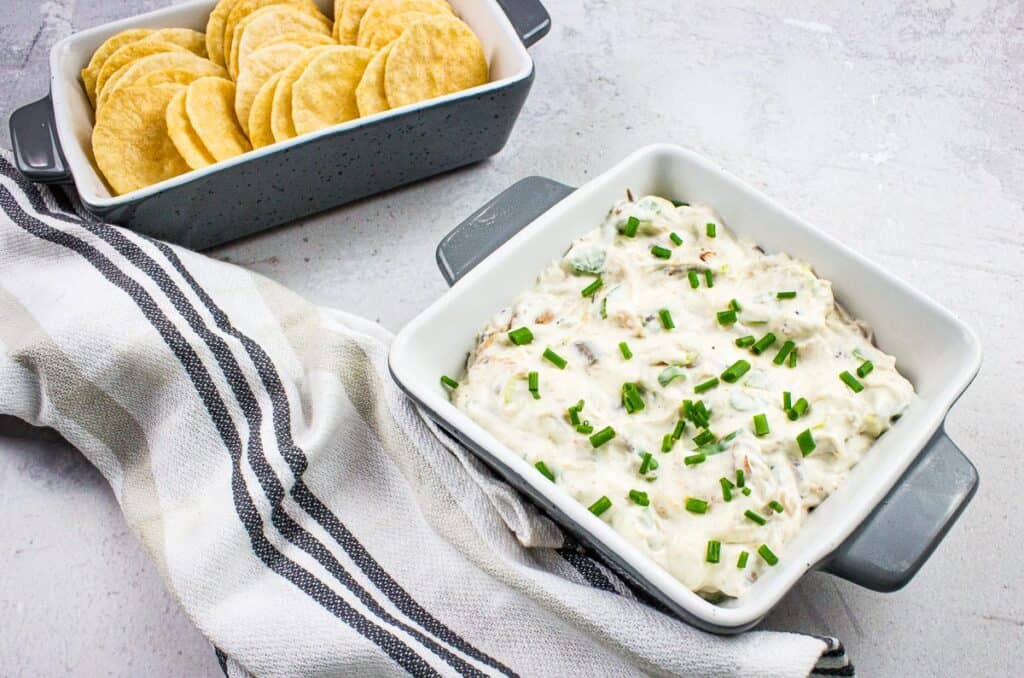 Two gray dishes: one with round crackers, the other with creamy dip topped with chopped green onions, placed on a striped cloth.
