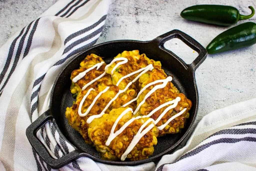 Corn fritters drizzled with cream in a black skillet, placed on a striped cloth, with two whole green jalape&ntilde;os next to it.
