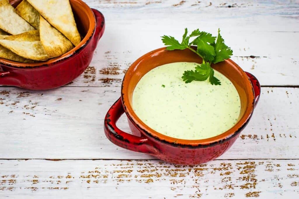 A red bowl filled with green sauce garnished with cilantro, placed on a rustic wooden table. Tortilla chips are in a separate small red dish beside it.