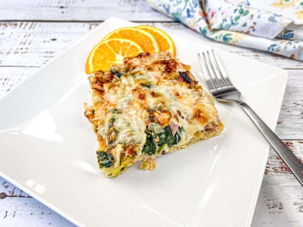 Slice of baked dish with spinach and cheese on a square white plate, garnished with two orange slices, beside a fork.