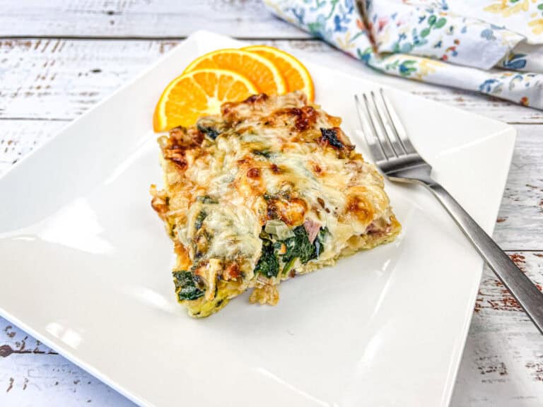 Slice of baked dish with spinach and cheese on a square white plate, garnished with two orange slices, beside a fork.