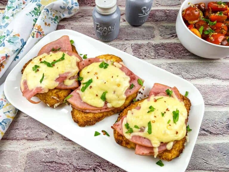Three pieces of ham and cheese toast sit on a white plate, garnished with herbs. A bowl of cherry tomato salad and salt and pepper shakers are in the background.