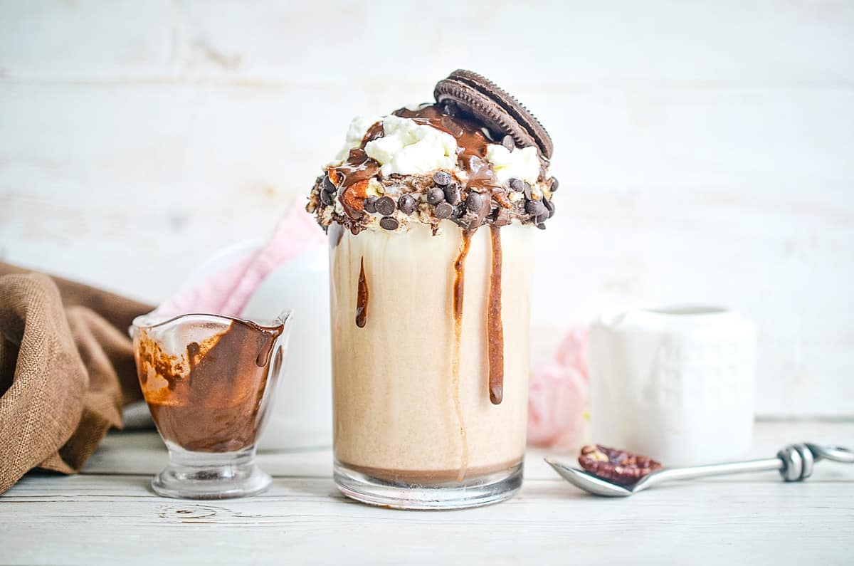 Tall glass of cookie dough milkshake surrounded by a small container of chocolate sauce.
