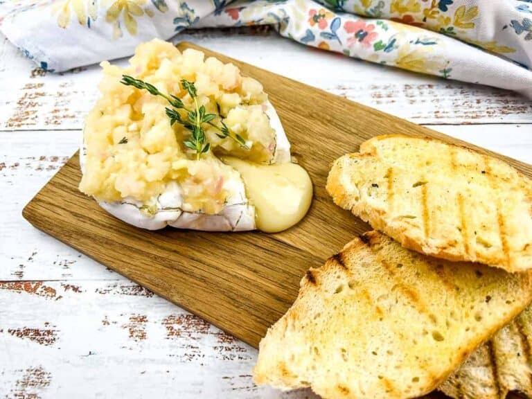 Baked brie topped with apple mixture and thyme on a wooden board, served with grilled bread slices.