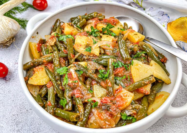 A bowl of cooked green beans, potatoes, and tomatoes garnished with parsley, with a spoon placed inside.