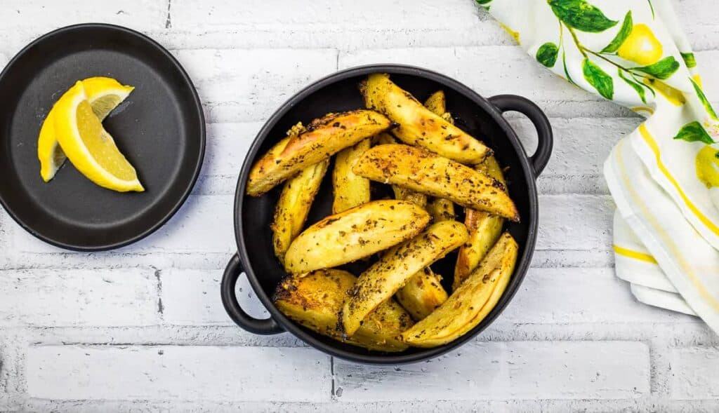 Potato wedges seasoned with herbs in a black bowl; lemon wedges on a small black plate beside a lemon-patterned cloth on a white brick surface.