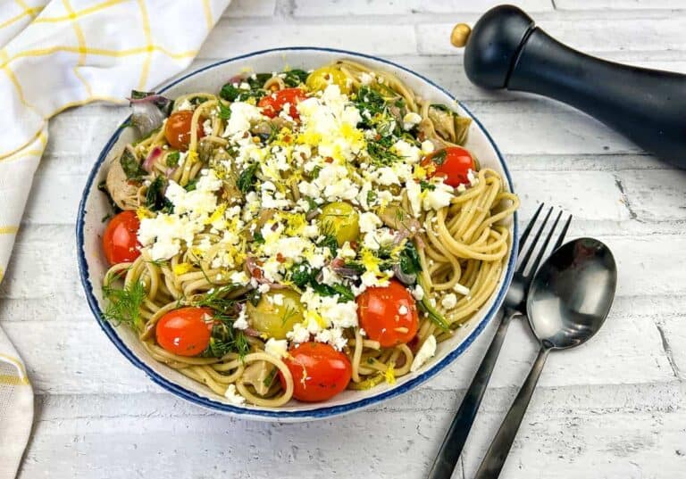A bowl of pasta topped with cherry tomatoes, greens, crumbled cheese, and a yellow garnish, with a fork, spoon, and pepper grinder nearby on a white surface.