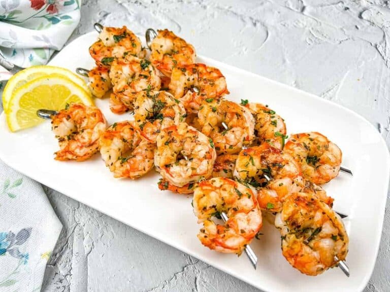Grilled shrimp skewers on a white plate, garnished with herbs, and served with lemon wedges.
